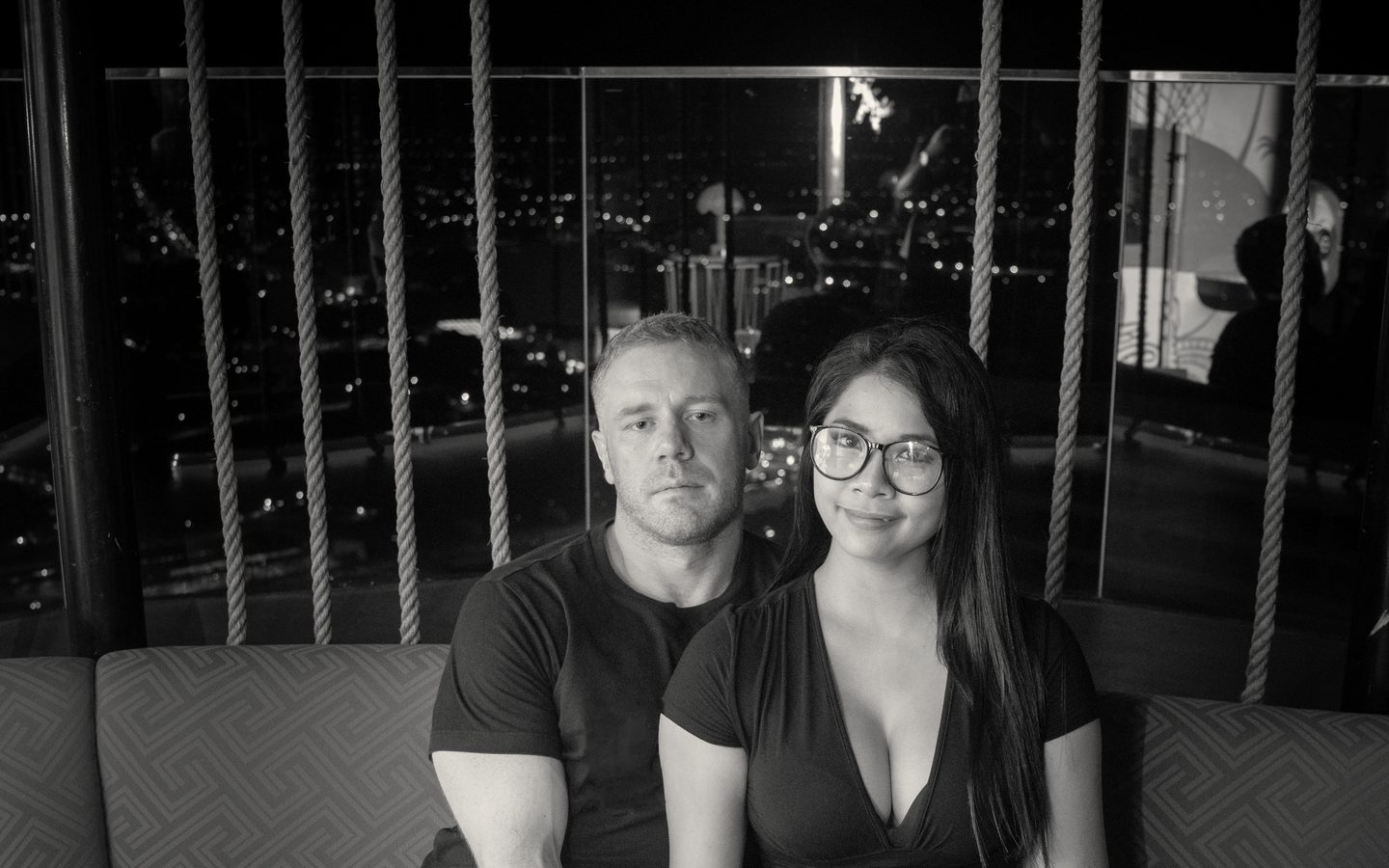 Tim and Paemi seated side by side on a rooftop sofa above Pattaya at night, facing the camera